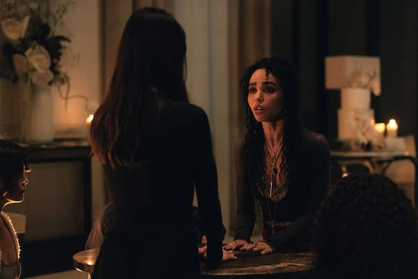 A dramatic scene featuring two women engaged in a tense conversation at a candlelit table, with one woman expressing concern while the other listens intently. Background elements include dim lighting and soft furnishings, enhancing the intimate and mysterious atmosphere.