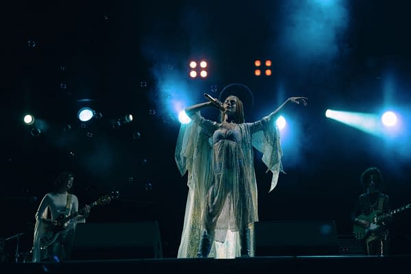 A performer in a flowing, ethereal gown sings passionately on stage, surrounded by soft lighting and floating bubbles, with a guitarist and another musician in the background.