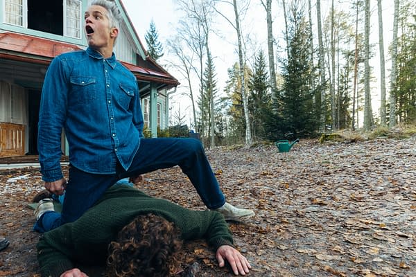 A man in a denim shirt poses dramatically over another person lying face down on the ground, surrounded by a wooded area. The scene suggests a tense or humorous moment, potentially linked to the title 'Over Your Dead Body.'