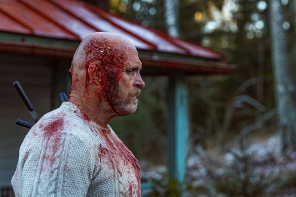 A man with a bald head and a mustache stands outdoors, covered in blood and wearing a white sweater. Blood drips from his head, and several knives are visibly stuck in his back, creating a grim atmosphere.