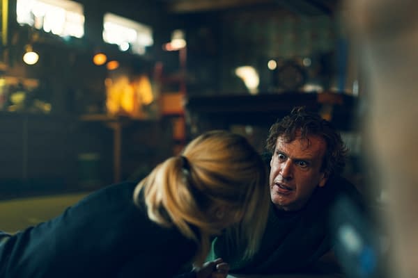 A tense moment from the film 'Over Your Dead Body,' featuring a man with wild hair and an intense expression, engaging in a dramatic scene with a woman who is leaning closely towards him. The background is dimly lit with hints of a cluttered workspace.