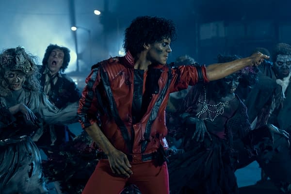 Jaafar Jackson portrays Michael Jackson in a dynamic scene, wearing a distinctive red jacket with black details, surrounded by a group of zombie-like figures. The atmosphere is moody and dark, reflecting a dramatic moment from the upcoming biopic 'Michael'.