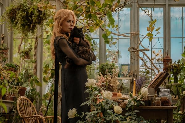 A woman with long blonde hair, dressed in a black gown, holds a fluffy black cat while standing in a greenhouse filled with lush greenery and white roses. A wooden table in front of her is adorned with candles and various botanical items, creating a mystical atmosphere.