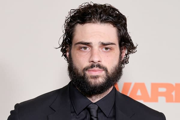 A man with a full beard and curly hair, wearing a black suit and tie, looks seriously at the camera against a neutral background with the word 'WARNER' visible.