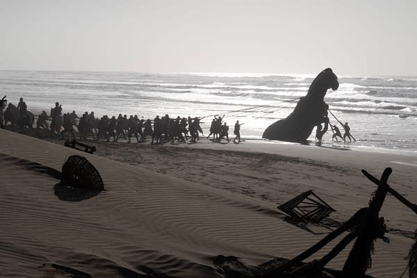 A dramatic beach scene from The Odyssey shows a group of people pulling a giant horse-shaped figure toward the shore through the surf.
