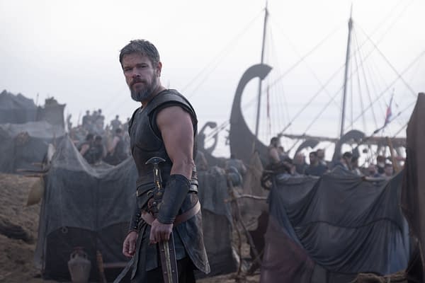 Matt Damon as Odysseus stands in armor at a crowded ancient Greek encampment beside a ship.
