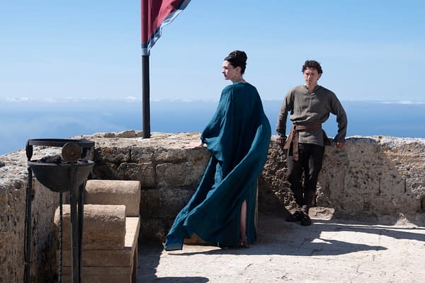 Anne Hathaway and Tom Holland stand on a stone terrace overlooking the sea in a scene from The Odyssey, with a tall flagpole and blue sky behind them.