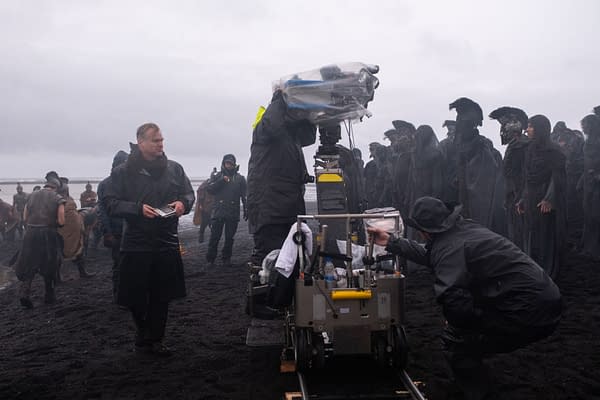 Christopher Nolan stands on a foggy film set beside a camera crew in dark rain gear while filming The Odyssey.