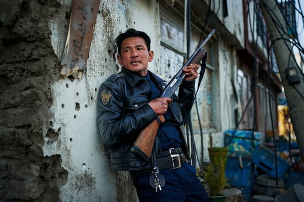 A tense film still shows a police officer in a leather jacket holding a shotgun while leaning against a damaged wall in a narrow, cluttered alley.