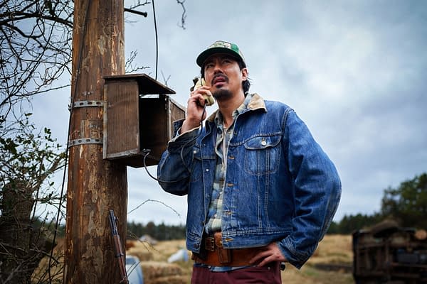 A man in a denim jacket and cap speaks on a vintage phone beside a weathered wooden utility pole in a rural field, from Hope.