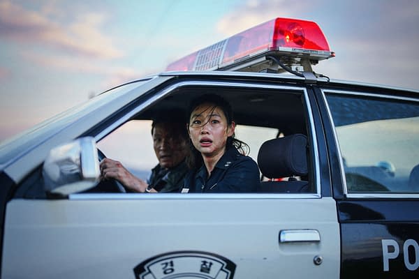 A tense scene in a police car with a frightened woman in uniform looking out the open window, while another officer drives. Red emergency lights glow on the roof against a cloudy sky.