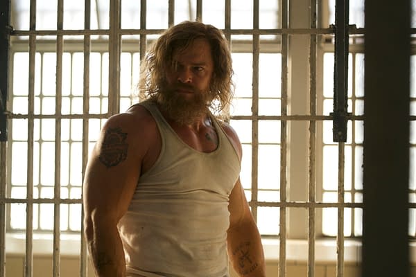 A rugged man with long hair and a beard stands in a white tank top behind metal bars, lit by bright windows in Motor City.