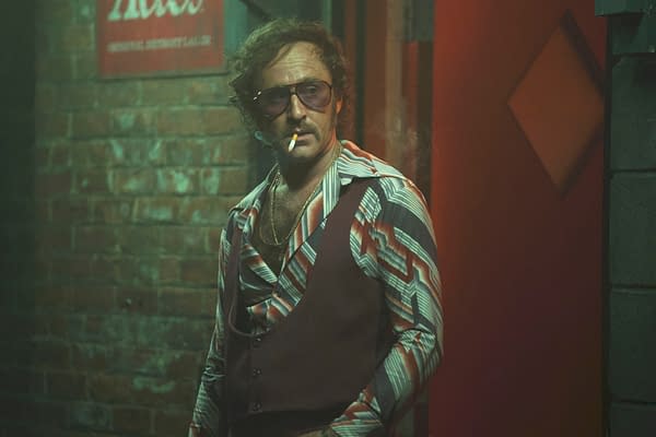 A man in sunglasses and a retro patterned shirt stands smoking against a brick wall in a moody scene from Motor City.