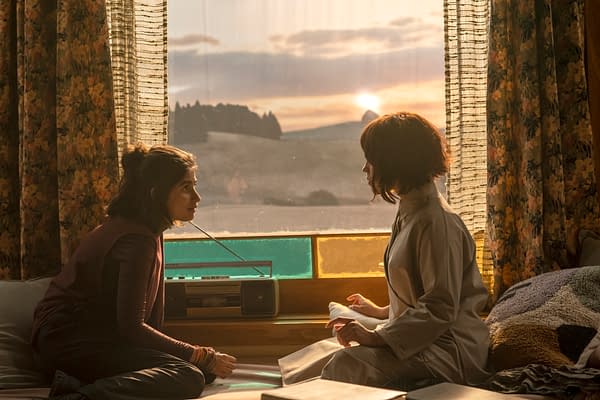 Two young women sit facing each other by a window at sunset, with a radio on the sill and patterned curtains framing a pastoral landscape outside. The warm, cinematic scene is from Klara and the Sun.