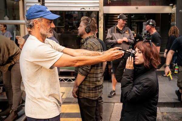A man adjusts a vintage camera while a woman in a black coat poses with her eye to the viewfinder on a film set. Crew members stand blurred in the background.