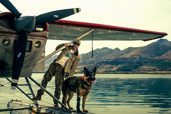 Brad Pitt stands on a floatplane with a German shepherd beside him, set against a calm lake and mountains in Heart of the Beast.