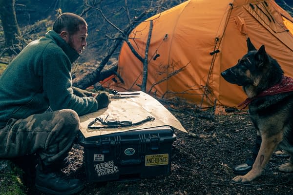 Brad Pitt as James Belmont kneels beside a German shepherd near an orange tent in a rugged forest camp in Heart of the Beast.