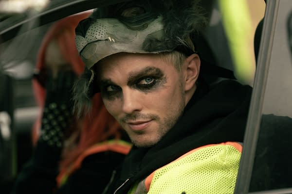 Nicholas Hoult as Ryan sits in a car wearing dramatic makeup and a worn animal mask in a scene from How to Rob a Bank.