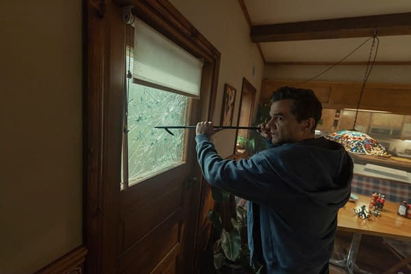 Wagner Moura as Jason in The Last House braces a crowbar against a shattered door window inside a warm, wood-paneled home.