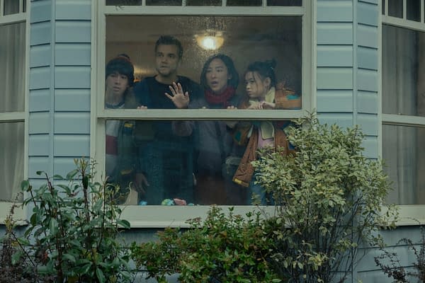 Four people stand pressed against a rain-speckled window inside a suburban house, looking out anxiously in The Last House.