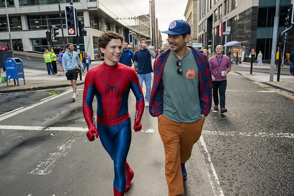 Spider-Man actor Tom Holland walks down a city street with a crew member during filming, smiling in his red-and-blue suit.