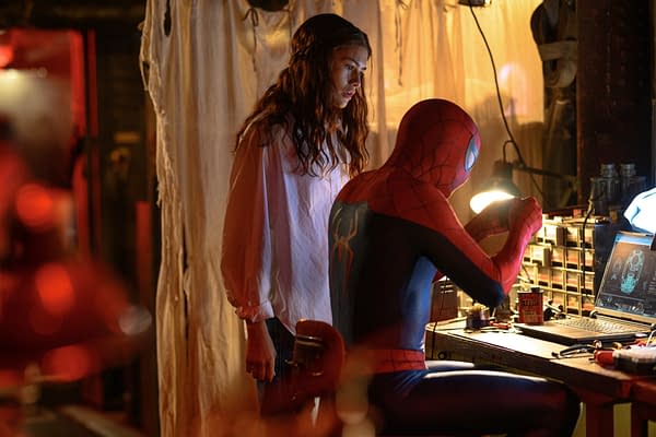 Spider-Man in costume works at a cluttered desk while a young woman watches nearby in a dimly lit workshop.