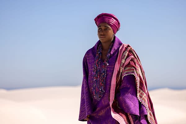 Lashana Lynch as Jumoke in Children of Blood and Bone, wearing a vivid purple and pink traditional outfit against a bright desert sky.