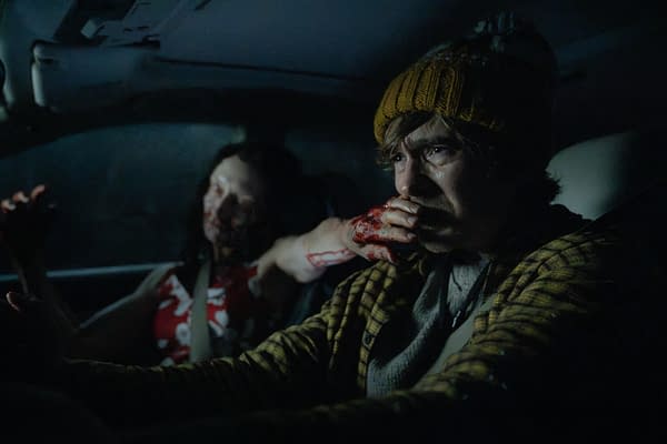 A tense Resident Evil scene inside a dark vehicle shows a man in a yellow knit hat biting a bloodied hand while a wounded figure sits behind him.