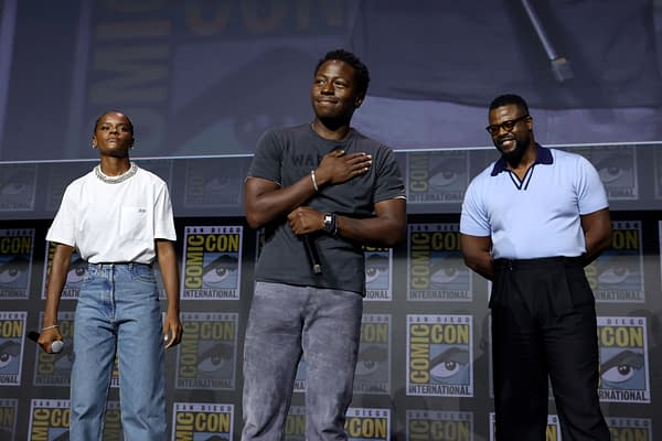 Letitia Wright, David Jonsson, and Winston Duke stand onstage at the Marvel Studios panel at San Diego Comic-Con, with Jonsson holding a microphone and smiling.