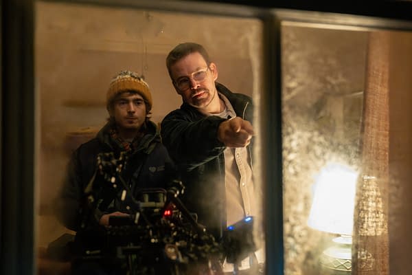 Two men peer through a window into a dimly lit room, one pointing ahead as if investigating something in a Resident Evil scene.