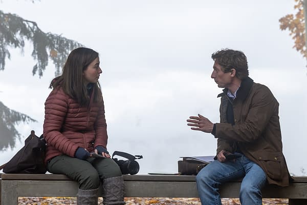 Two people sit on a bench outdoors in a foggy setting, talking intently. A camera rests between them, suggesting a film or documentary scene.
