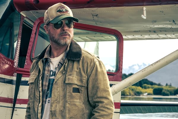 Brad Pitt stands beside a small airplane in a scene from Heart of the Beast, wearing a cap, aviator sunglasses, and a worn jacket.