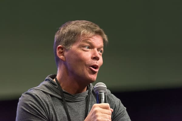 Rob Liefeld speaks into a handheld microphone at a comic convention panel in Los Angeles. He wears a gray hoodie and looks animated mid-sentence.