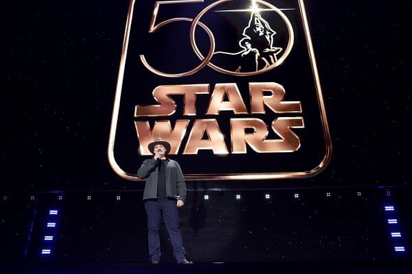Dave Filoni stands onstage at Disney Entertainment Showcase in front of a giant Star Wars 50th anniversary logo, speaking into a microphone.