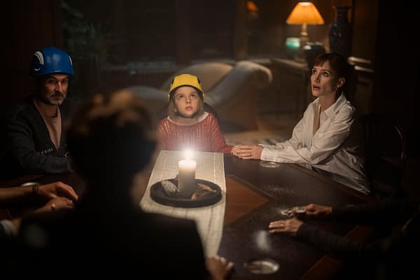 Jessica Chastain, Jay Duplass, and a young girl sit at a dimly lit table in Other Mommy, wearing colorful helmets and holding hands.