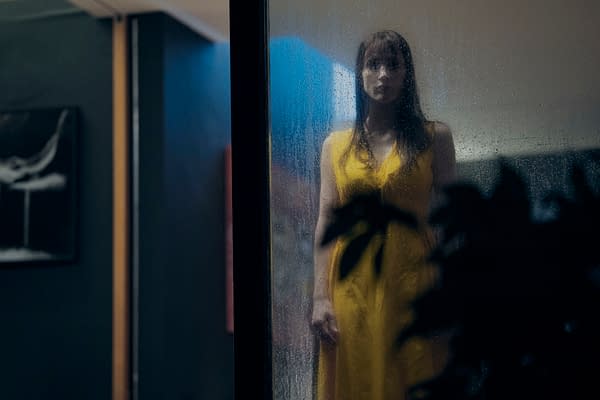 Jessica Chastain as Other Mommy stands behind a rain-speckled glass door in a yellow dress, in a tense scene from the horror film Other Mommy.
