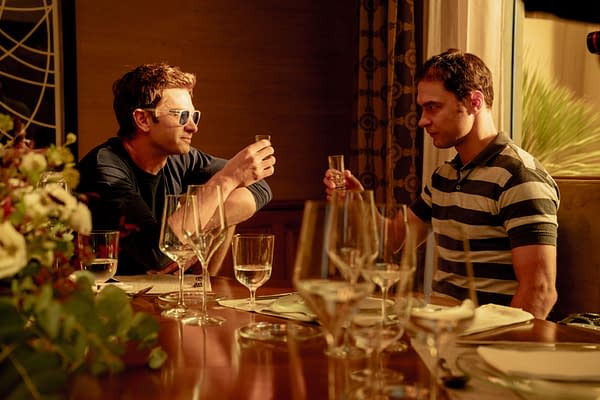 Andrew Garfield and Yura Borisov sit across a candlelit dinner table, raising small glasses in a tense conversation scene from Artificial.
