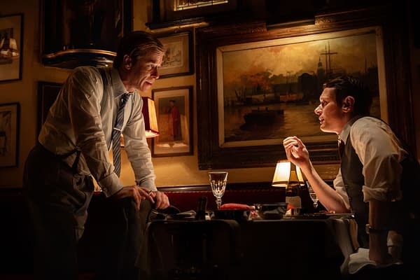 Guy Pearce and Jack O'Connell face each other in a dimly lit restaurant scene from INK, with warm lamps and framed paintings in the background.