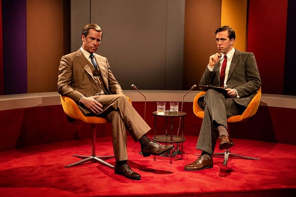 Guy Pearce as Murdoch sits in a retro TV studio interview set beside a host, both in suits and facing forward.