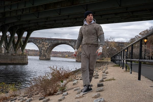 Anthony Ippolito, as Sylvester Stallone in I Play Rocky, walks along a riverside path beneath a bridge, wrapped in a gray hoodie and sweatpants with bandaged hands.