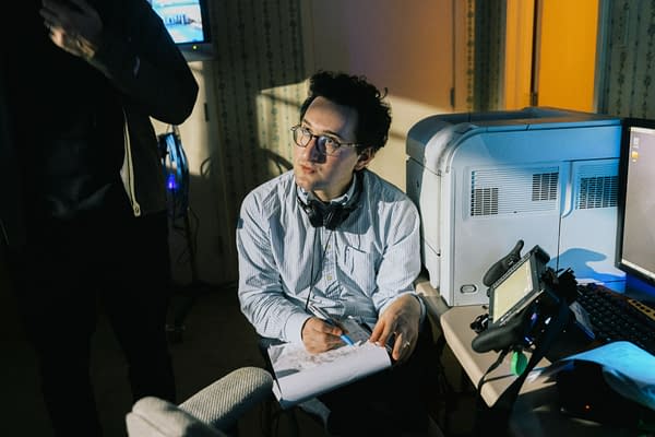 A man with glasses and headphones around his neck sits in a dimly lit office or studio, taking notes beside computer equipment and a large printer. The scene has a cinematic, behind-the-scenes feel.