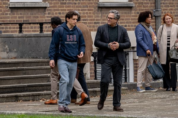 A young man in an ESU hoodie walks beside a gray-haired man in glasses outside a brick building, with several other people in the background.