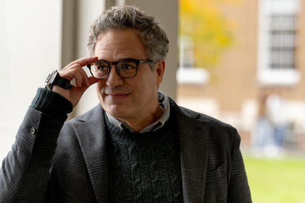 A middle-aged man with gray hair and glasses adjusts his frames while wearing a blazer indoors, with a softly blurred windowed background.