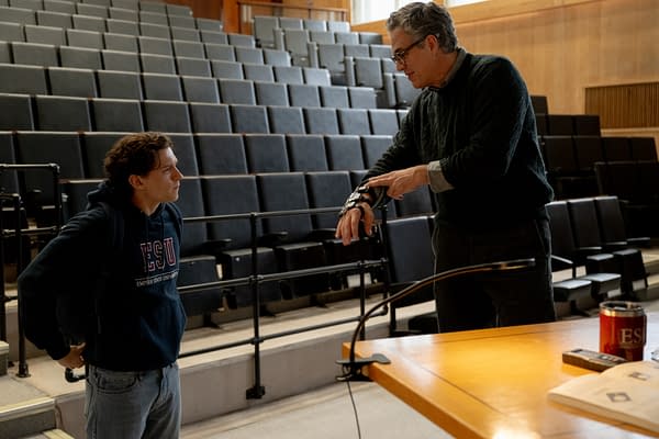 Two men confer in a lecture hall, one showing a wrist device or prosthetic arm to the other. Rows of empty auditorium seats rise behind them.