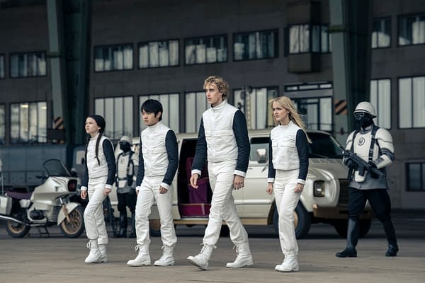Four young characters in white uniforms walk across an industrial set with armored guards and vehicles behind them in The Hunger Games: Sunrise on the Reaping.