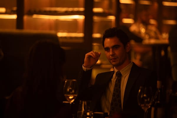 A man in a suit sits at a dimly lit restaurant table under warm amber lighting, looking across at someone off camera.