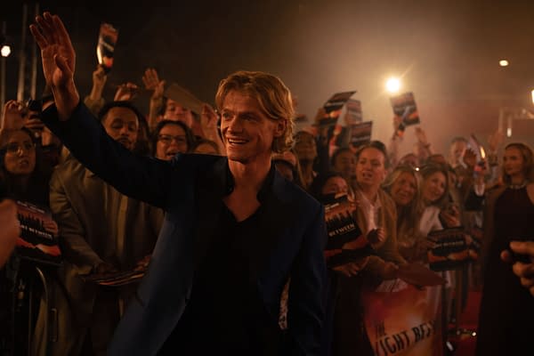Clayface stands smiling and waving to a cheering crowd at a crowded nighttime event, with fans holding posters and reaching for him.