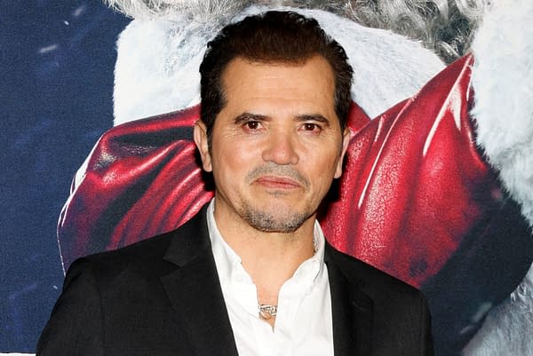 John Leguizamo poses on the red carpet at a movie premiere event in Hollywood, with a festive background featuring a Santa Claus image. He is dressed in a black suit and white shirt.