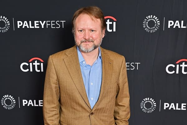 Rian Johnson, a film director, stands smiling at a red carpet event, wearing a brown suit and blue shirt, against a black backdrop featuring the PaleyFest logo.