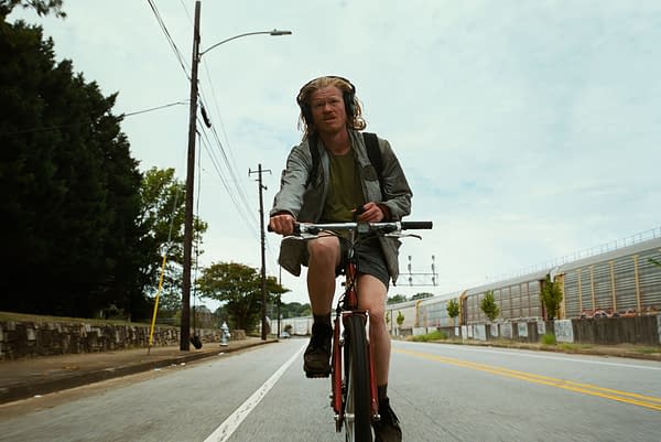 A scene from the film 'BUGONIA' (2025) featuring a man with long hair wearing headphones, riding a bicycle along a city street. He appears focused and is dressed in casual attire with a backpack.
